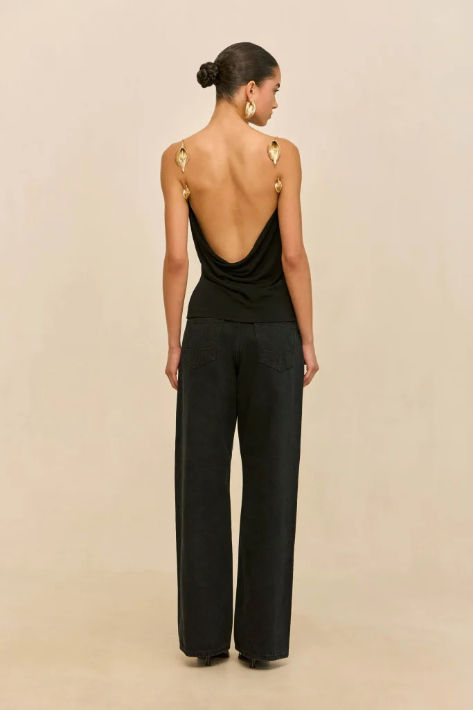 Back view of a woman wearing a black draped open-back top, gold leaf earrings, and wide-leg black pants.