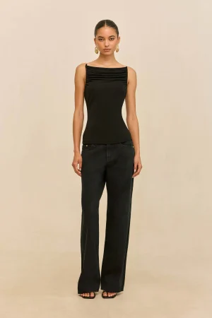 Full-body portrait of a model wearing a black sleeveless top with a gathered neckline and dark jeans, standing against a beige backdrop with gold earrings.
