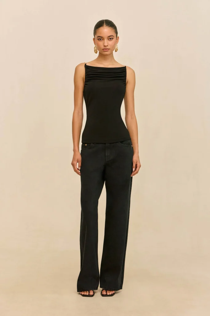 Full-body portrait of a model wearing a black sleeveless top with a gathered neckline and dark jeans, standing against a beige backdrop with gold earrings.
