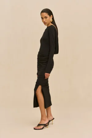 Side view of a model wearing a black long-sleeve dress with a thigh-high slit, gold hoop earrings, and black heeled sandals against a beige background.