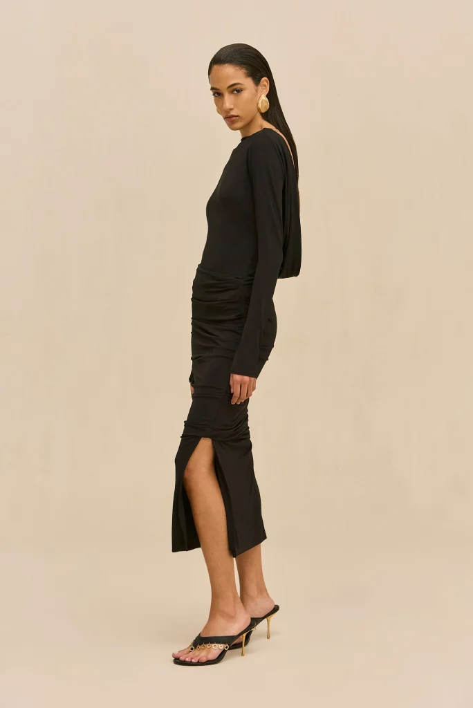 Side view of a model wearing a black long-sleeve dress with a thigh-high slit, gold hoop earrings, and black heeled sandals against a beige background.