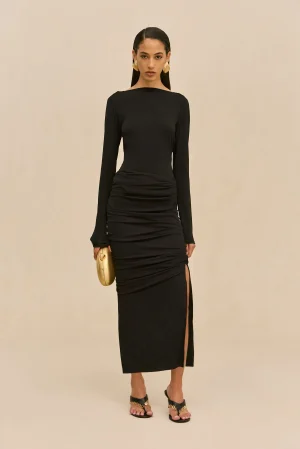 Woman in a fitted black long-sleeve dress with a high waist, holding a small gold clutch, standing against a neutral backdrop.