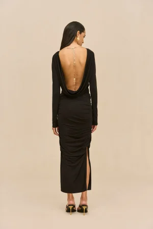 Back view of a woman in a black, backless dress with a gold chain running down her spine, standing against a beige backdrop, wearing heeled sandals.
