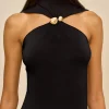 Close-up of a woman wearing a black halter-neck top with a gold knot detail at the center, and gold earrings against a beige background.