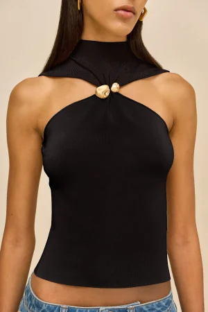 Close-up of a woman wearing a black halter-neck top with a gold knot detail at the center, and gold earrings against a beige background.