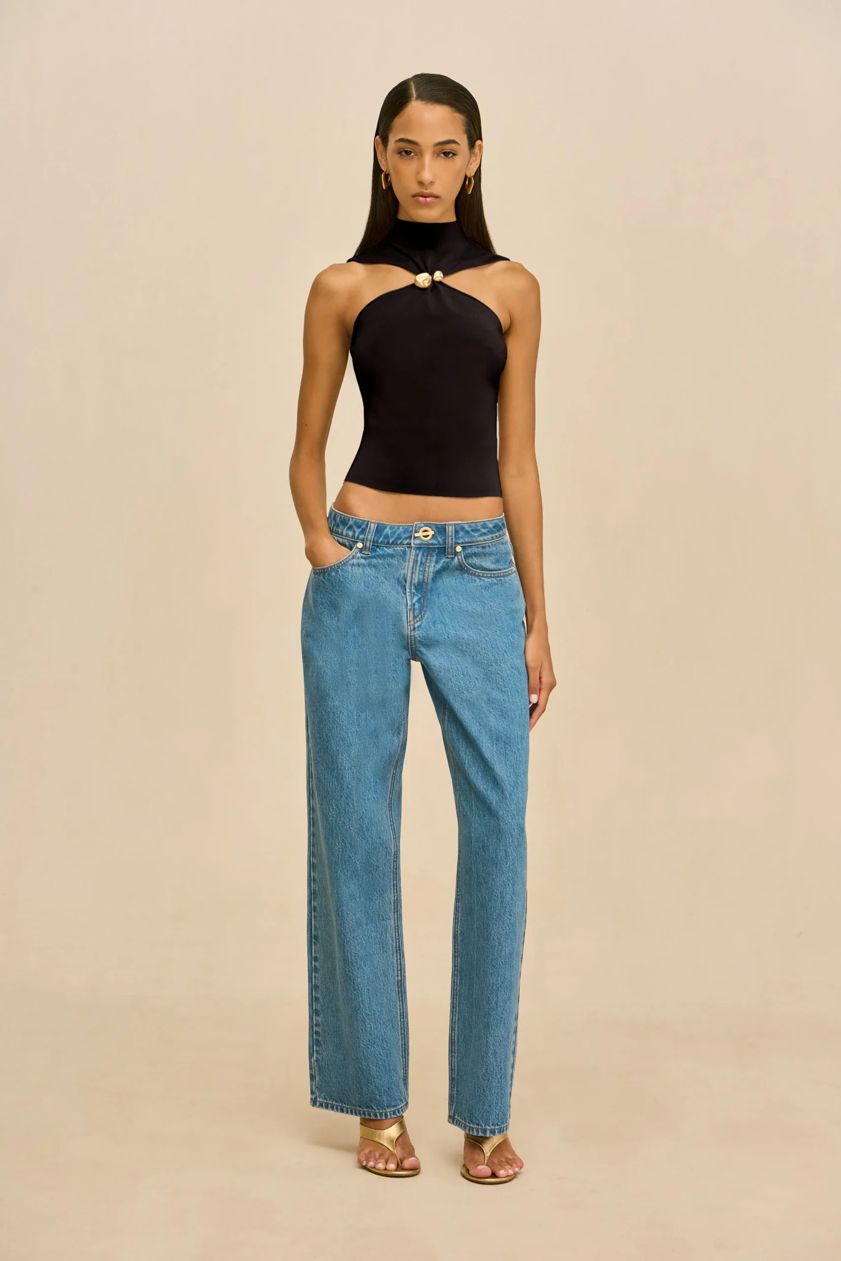 IMG_2561 - New Trend Woman wearing a black halter-style top with a knotted front detail and light-washed blue jeans, standing against a beige background.