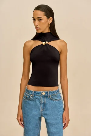 Model wearing a black halter-style top with a gold knot accent at the chest, paired with blue jeans, against a beige background.
