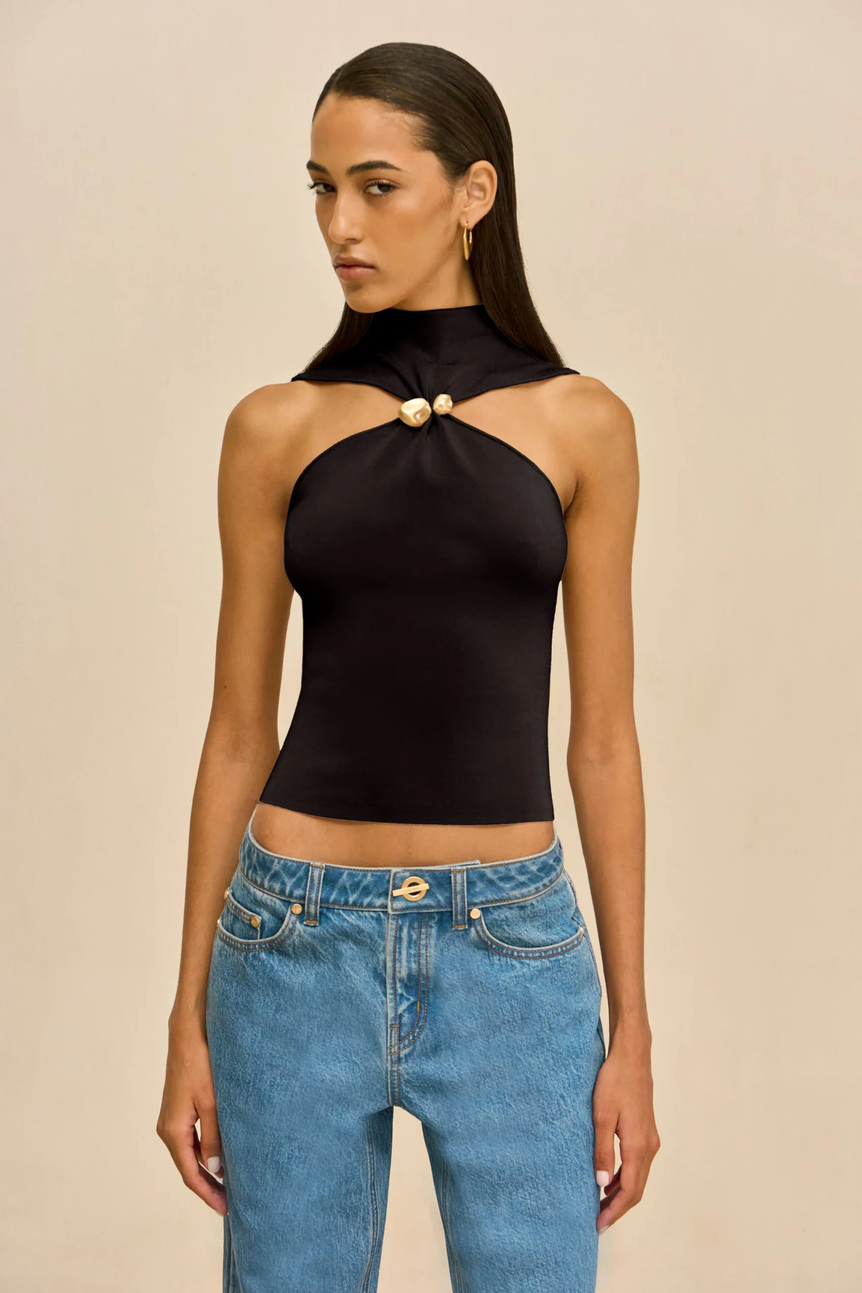 IMG_2562 - New Trend Model wearing a black halter-style top with a gold knot accent at the chest, paired with blue jeans, against a beige background.