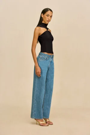 Woman in a black halter top and high-waisted blue jeans, standing in profile against a beige background.