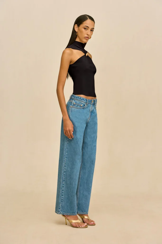 Woman in a black halter top and high-waisted blue jeans, standing in profile against a beige background.