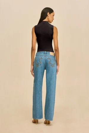 Back view of a woman in a black sleeveless top and blue jeans with decorative back pockets, wearing yellow high-heeled sandals against a beige background.