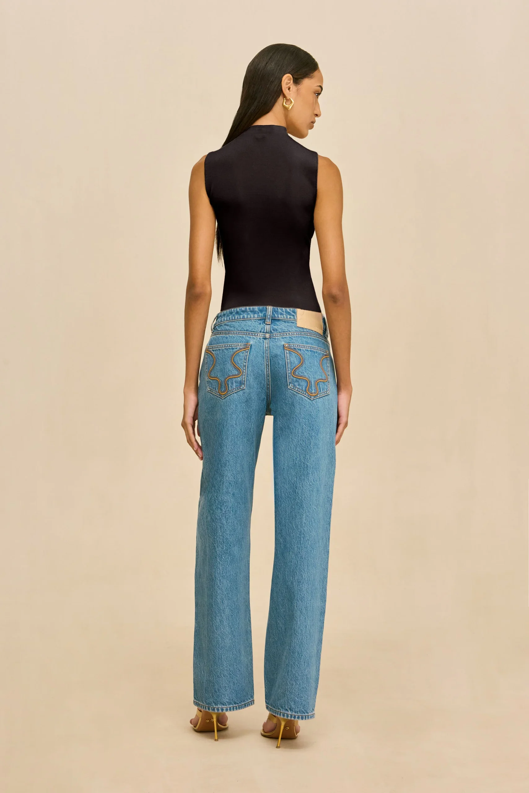 IMG_2564 - New Trend Back view of a woman in a black sleeveless top and blue jeans with decorative back pockets, wearing yellow high-heeled sandals against a beige background.