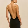 Back view of a woman in a black open-back dress with gold leaf shoulder straps and matching gold earring, hair in a sleek bun, beige background.