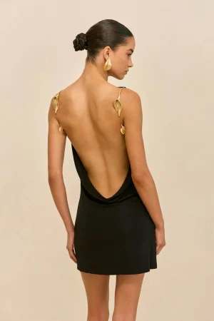Back view of a woman in a black open-back dress with gold leaf shoulder straps and matching gold earring, hair in a sleek bun, beige background.