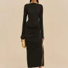Model wearing a black long-sleeve midi dress with ruching, holding a gold clutch and wearing gold hoop earrings, standing against a beige backdrop.