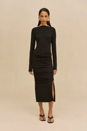 Woman in a sleek black long-sleeve midi dress with a side slit, standing against a beige backdrop, wearing gold earrings and sandals.