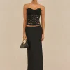 Woman in a strapless black lace top and floor-length black skirt, holding a small black handbag with a gold chain, against a beige backdrop.