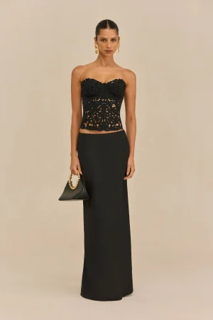 Woman in a strapless black lace top and floor-length black skirt, holding a small black handbag with a gold chain, against a beige backdrop.