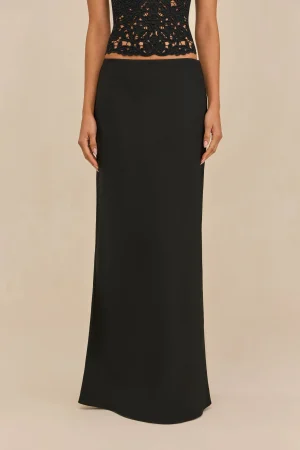 Person wearing a black lace crop top and a long black skirt, standing against a beige background.