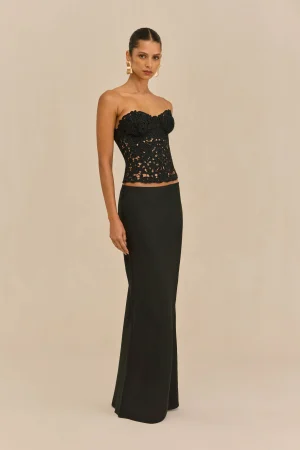 Model wearing a strapless black lace top with a long black skirt, gold statement earrings, against a beige backdrop.