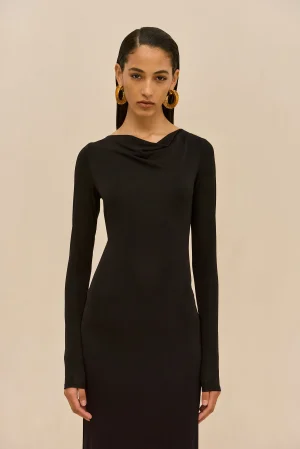 Woman in a black long-sleeve dress with oversized gold hoop earrings, standing against a beige wall.