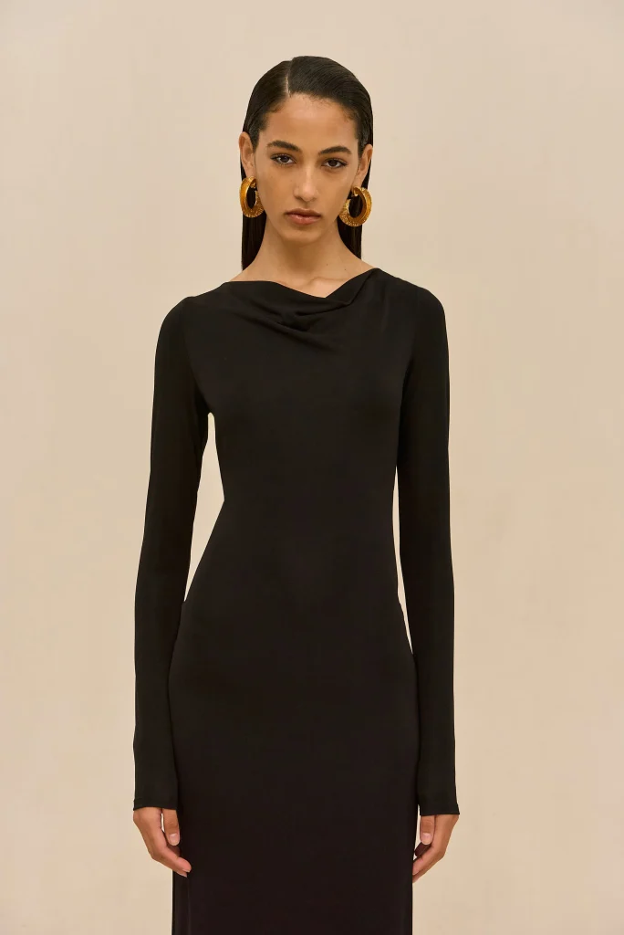 Woman in a black long-sleeve dress with oversized gold hoop earrings, standing against a beige wall.