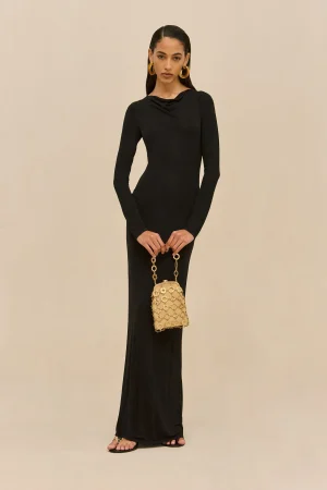 Full-length photograph of a woman in a black long-sleeve dress standing against a beige background, holding a gold chain handbag with a circular pattern.