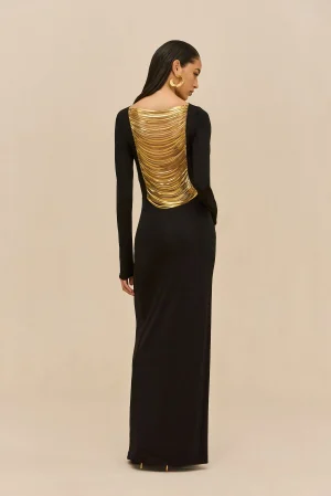 Woman in a long black gown seen from behind, wearing a gold multi-layered back accessory and large gold hoop earrings, against a beige backdrop.
