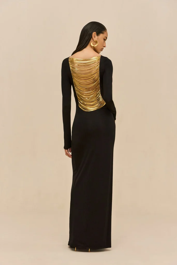 Woman in a long black gown seen from behind, wearing a gold multi-layered back accessory and large gold hoop earrings, against a beige backdrop.