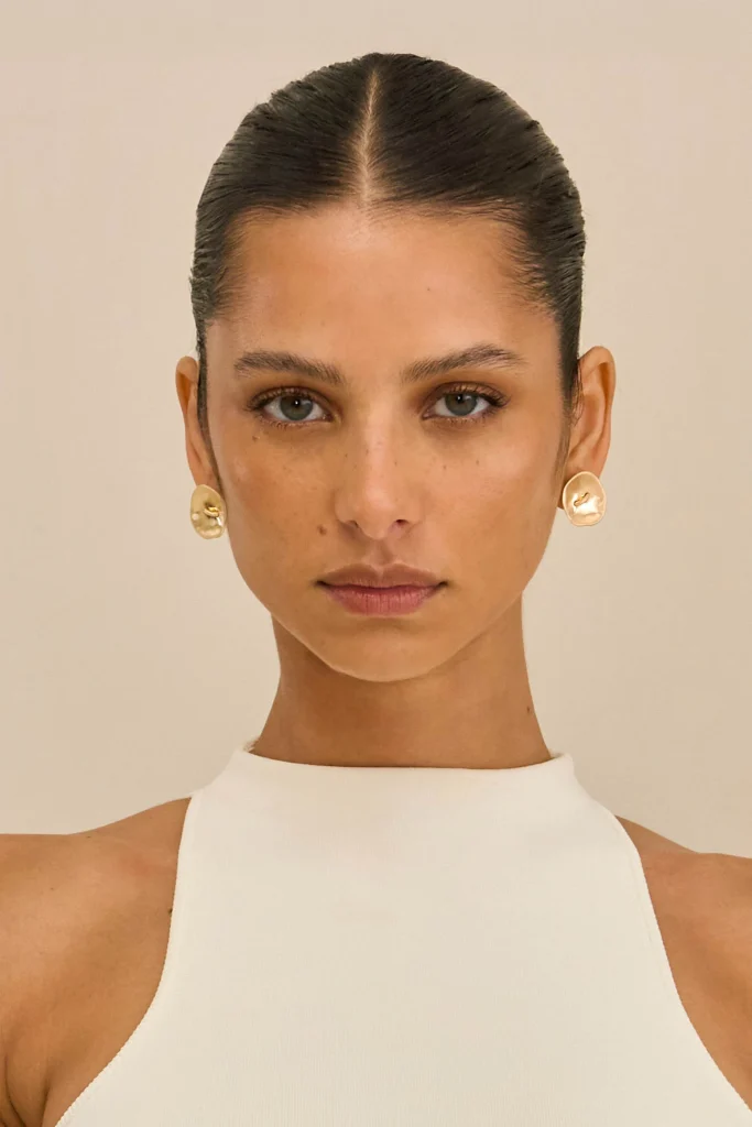 Woman with slicked-back hair, wearing a white sleeveless top and gold earrings, facing camera against neutral background.
