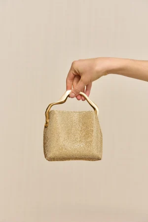 Hand holding a small gold beaded evening bag by its curved metal handle against a beige background.