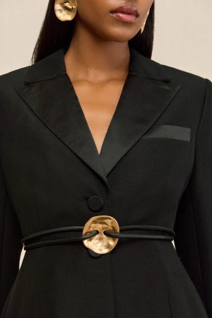 Close-up of a woman wearing a black blazer with wide satin lapels, a circular gold belt buckle, and large gold earrings visible near the ear.