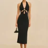 IMG_2614 - New Trend Full-body shot of a woman in a black halter-neck dress with waist cutouts, gold earrings, and strappy heeled sandals, holding a small black handbag.