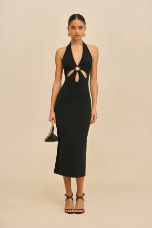 Full-body shot of a woman in a black halter-neck dress with waist cutouts, gold earrings, and strappy heeled sandals, holding a small black handbag.
