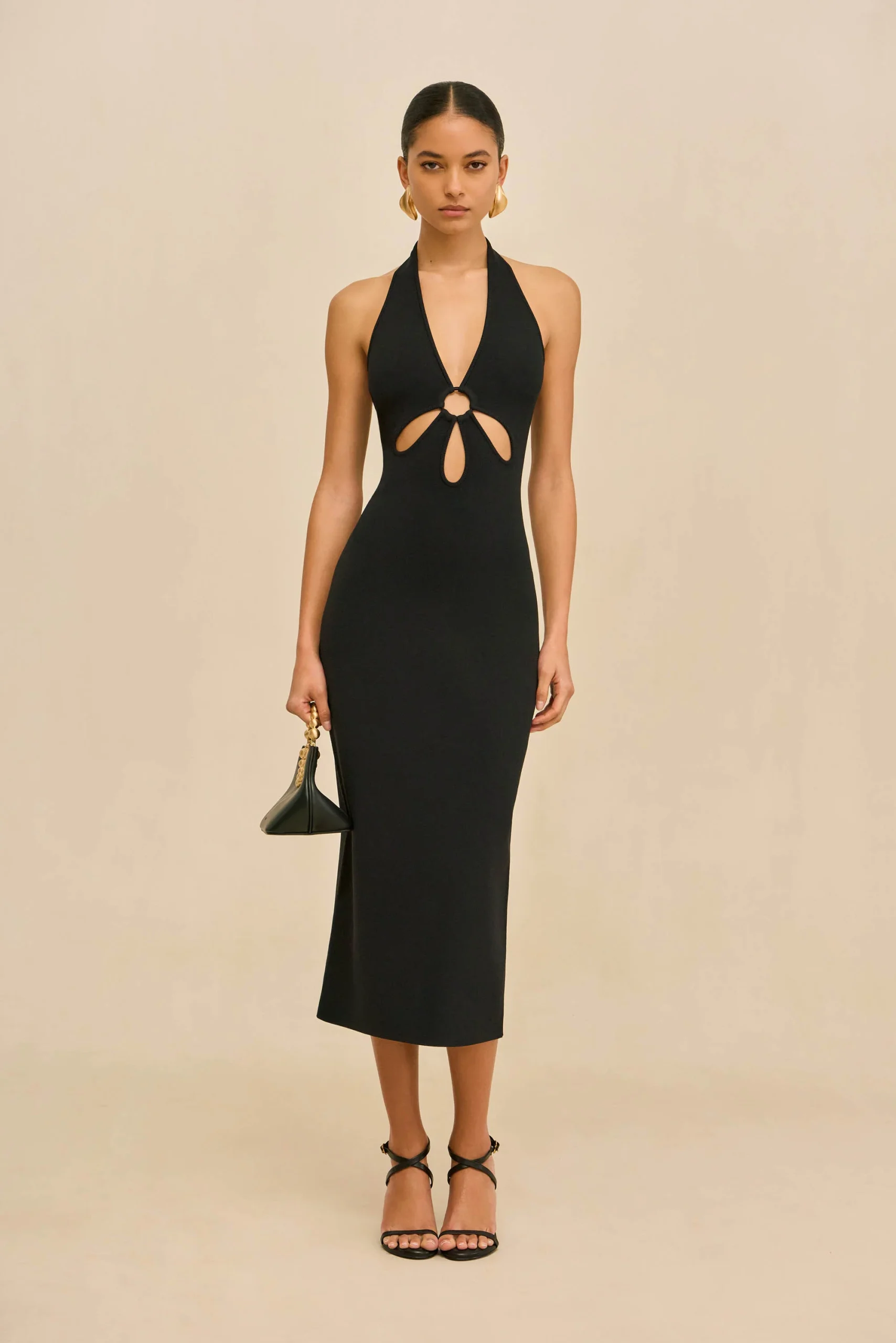 IMG_2614 - New Trend Full-body shot of a woman in a black halter-neck dress with waist cutouts, gold earrings, and strappy heeled sandals, holding a small black handbag.