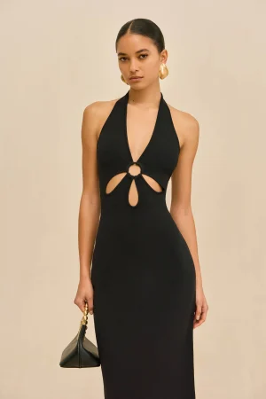 Woman wearing a black halter dress with front keyhole cutouts, large gold earrings, and a handbag against a beige background.
