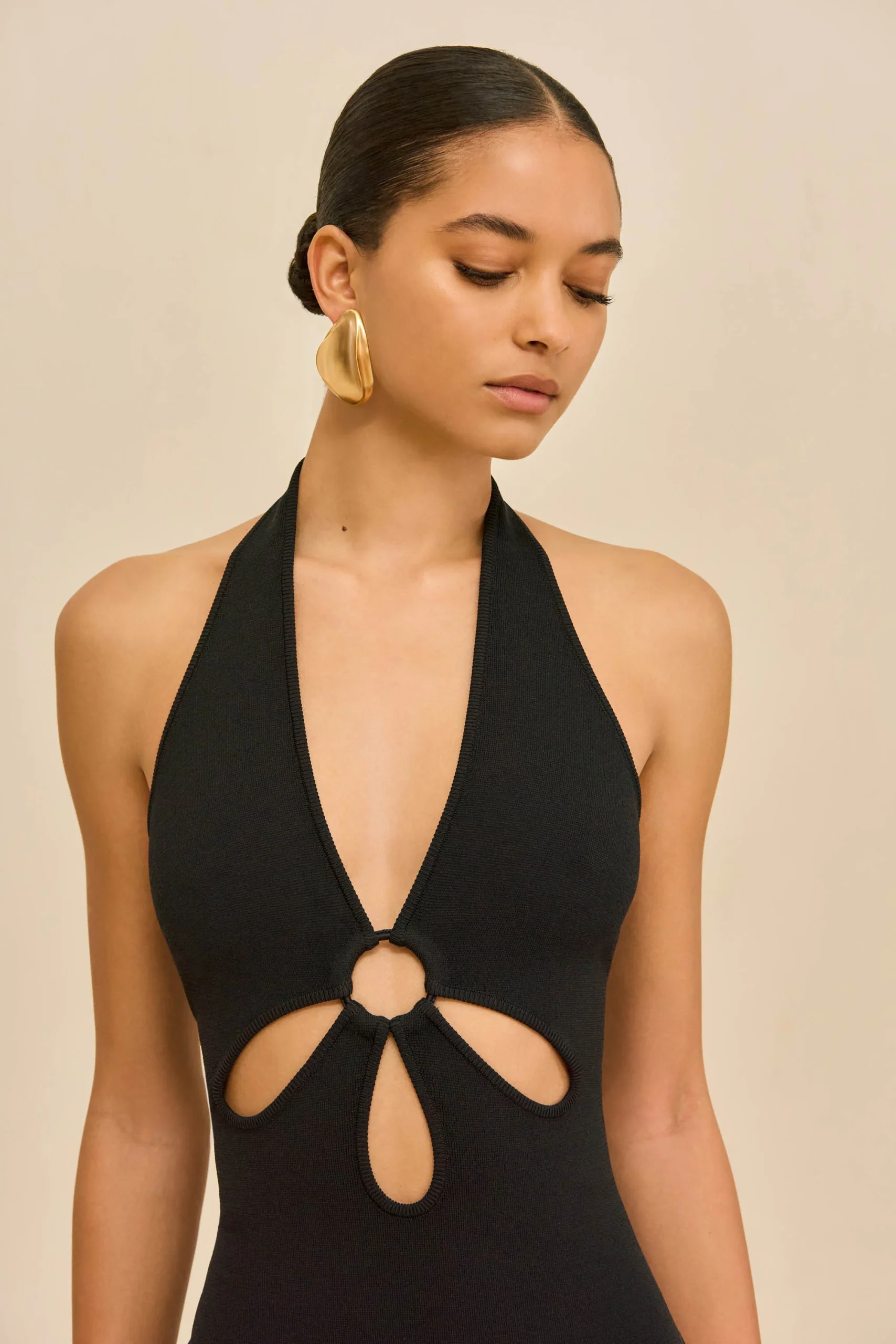 IMG_2616 - New Trend Model wearing a black halter swimsuit with large circular cutouts and a gold statement earring, against a beige background, head tilted downward.