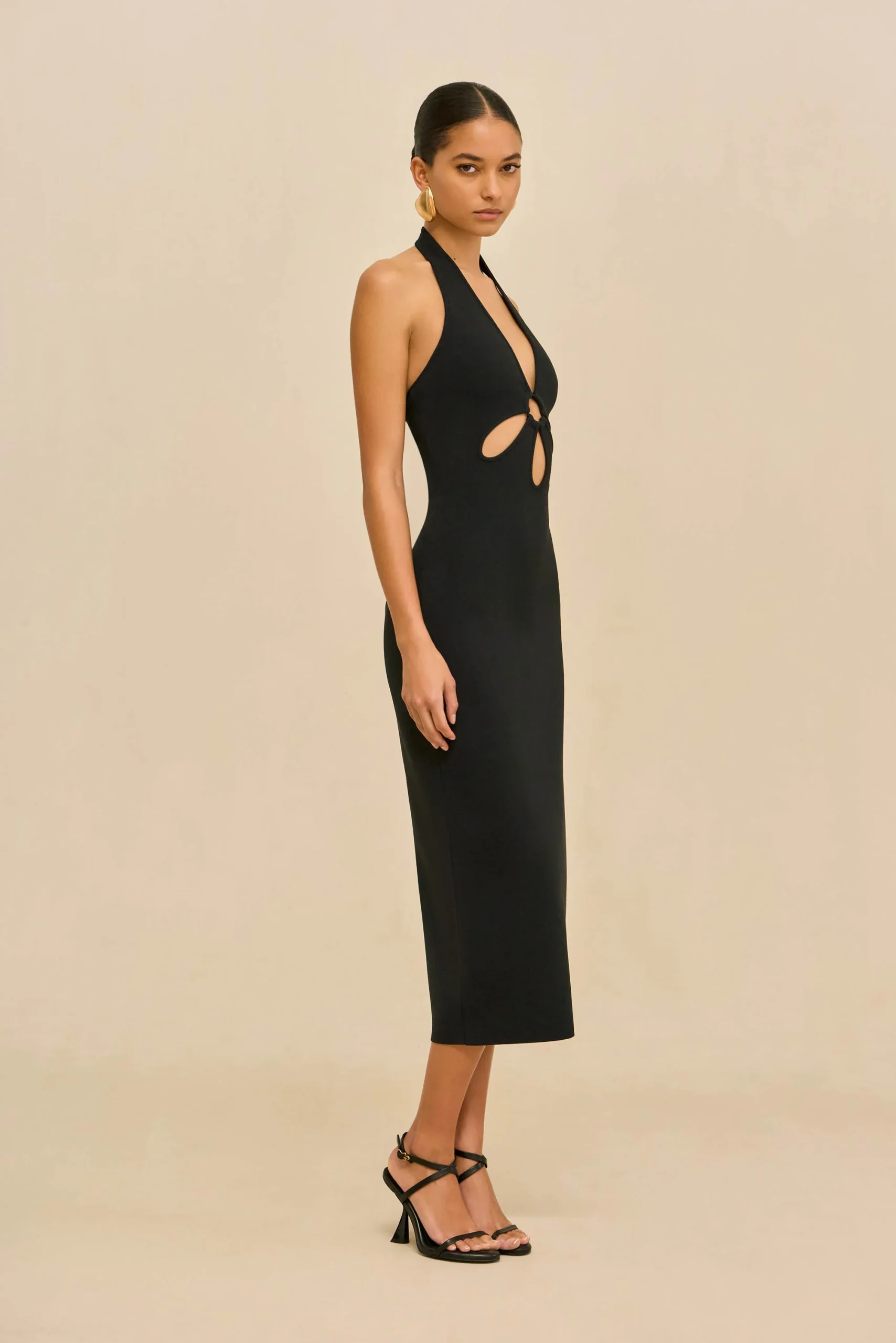 IMG_2617 - New Trend Model in a sleek black halter-neck dress with cutouts, standing sideways against a beige backdrop with strappy black heels and gold earrings.