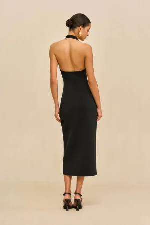 Back view of a woman in a black halter-neck dress with her hair in a bun, wearing gold earrings, standing in a neutral beige backdrop.