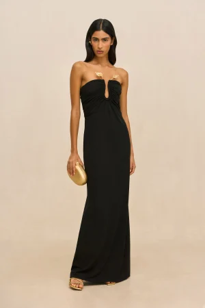 Full-length portrait of a woman in a black strapless gown with ruched center, gold leaf accents at the bust, holding a gold clutch, and wearing gold sandals against a beige backdrop