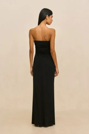 Back view of a woman wearing a floor-length black strapless dress, standing against a light beige backdrop, with straight hair.