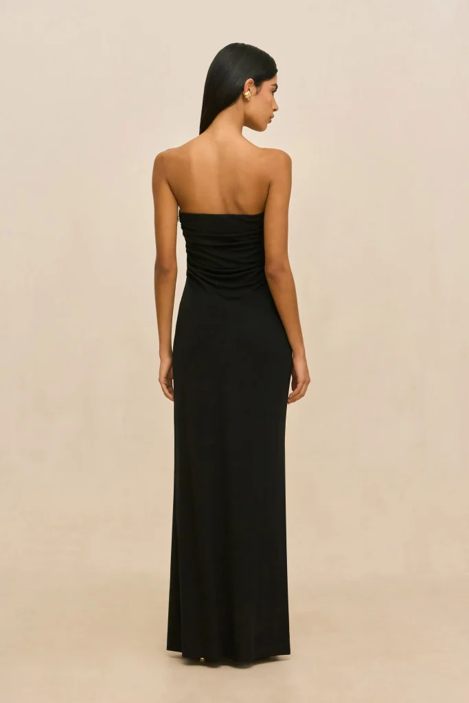 Back view of a woman wearing a floor-length black strapless dress, standing against a light beige backdrop, with straight hair.