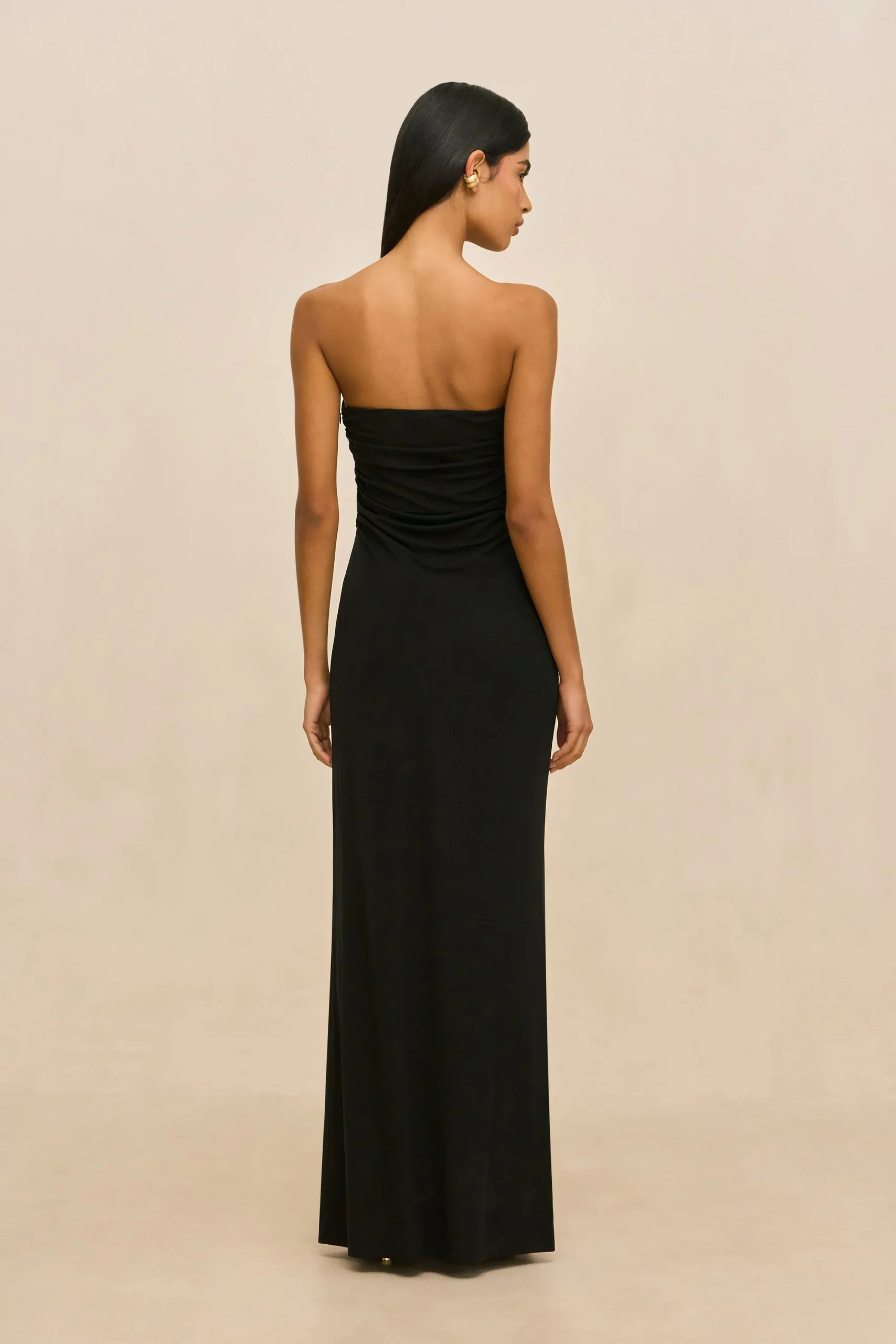 IMG_2624 - New Trend Back view of a woman wearing a floor-length black strapless dress, standing against a light beige backdrop, with straight hair.
