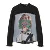 Black long-sleeve top with a large graphic print of a person’s portrait overlaid by green lace and flowers on the chest area
