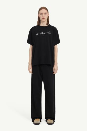 Male model wearing an oversized black T-shirt with a white handwritten logo, paired with wide-leg black pants and tan shoes against a light background.