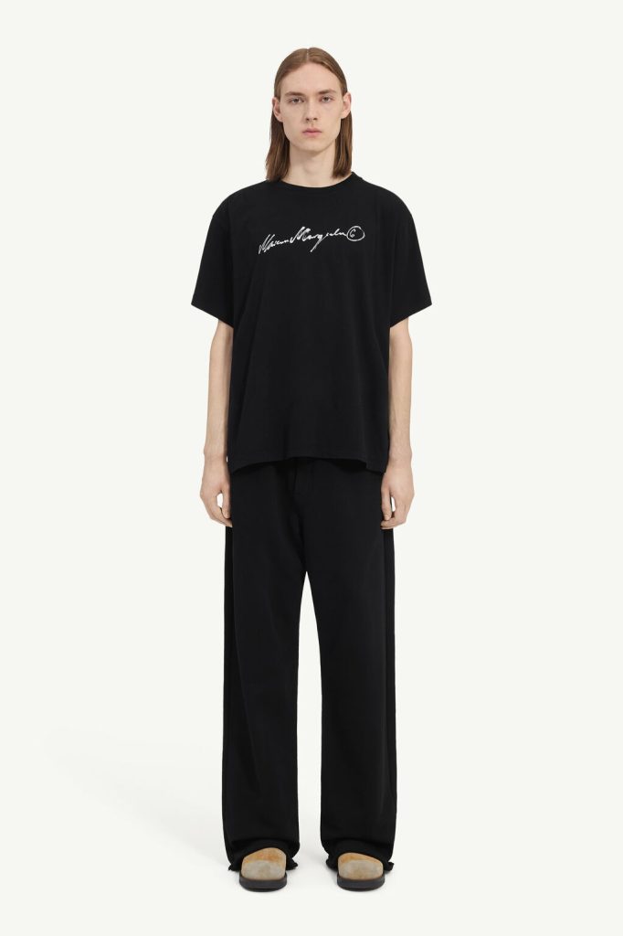 Male model wearing an oversized black T-shirt with a white handwritten logo, paired with wide-leg black pants and tan shoes against a light background.