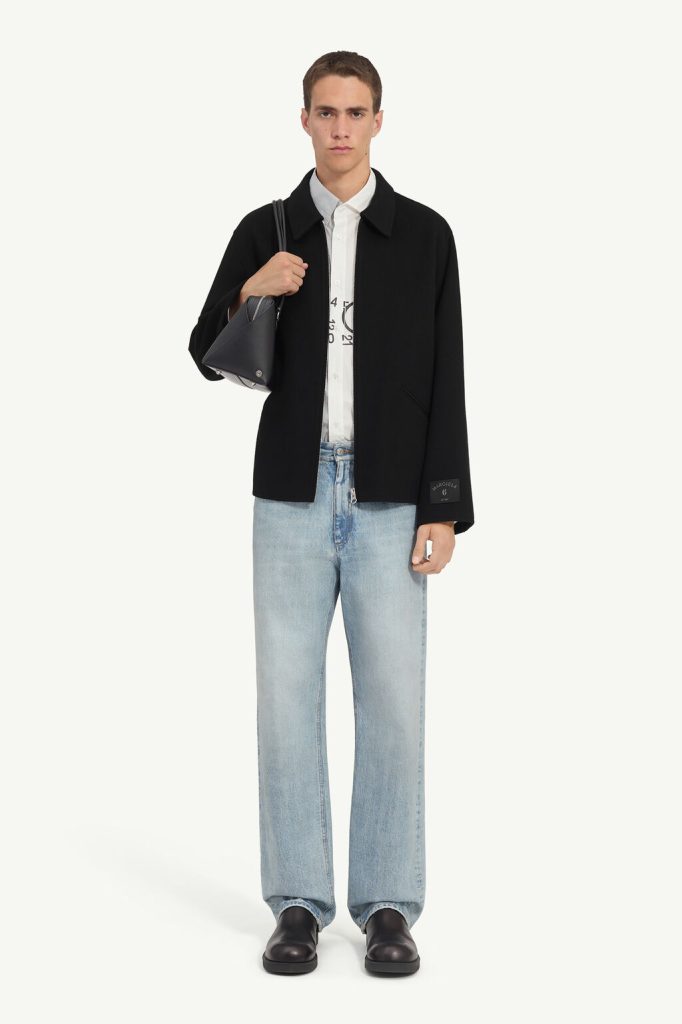 Male model wearing a black jacket over a white button-down shirt, light-wash jeans, black shoes, and carrying a black bag over the shoulder.