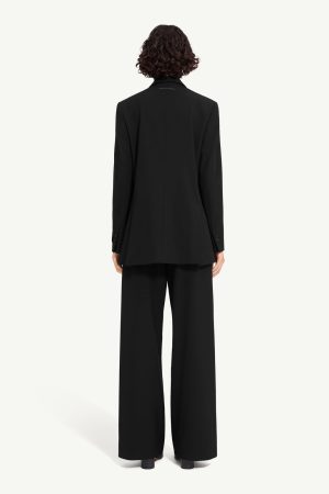 Person seen from behind wearing a black blazer and wide-leg trousers against a light backdrop, standing upright.