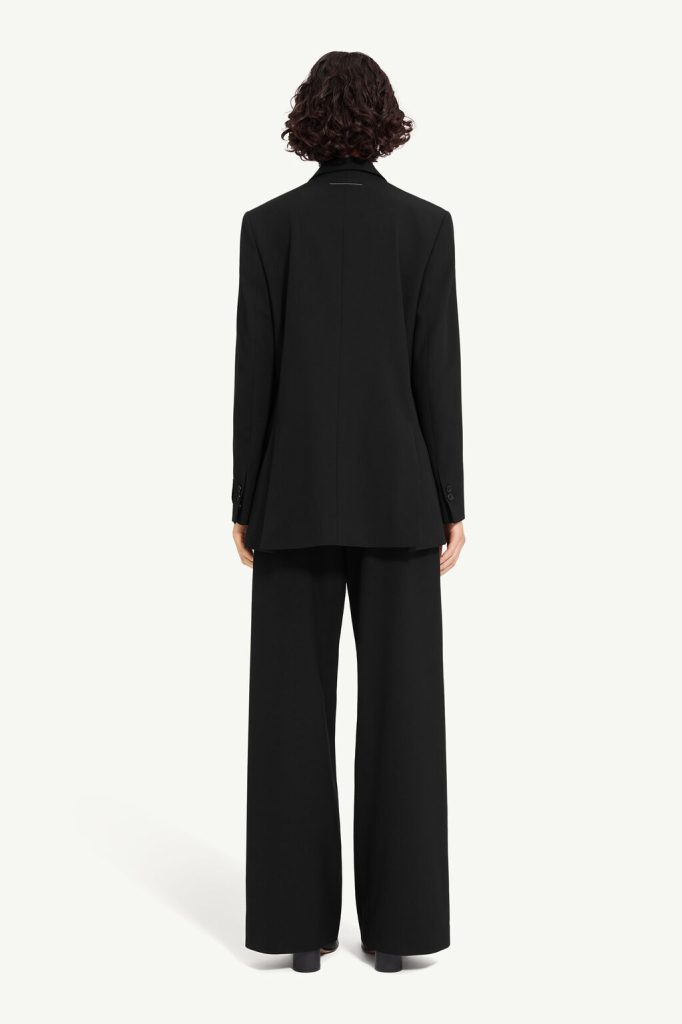 Person seen from behind wearing a black blazer and wide-leg trousers against a light backdrop, standing upright.