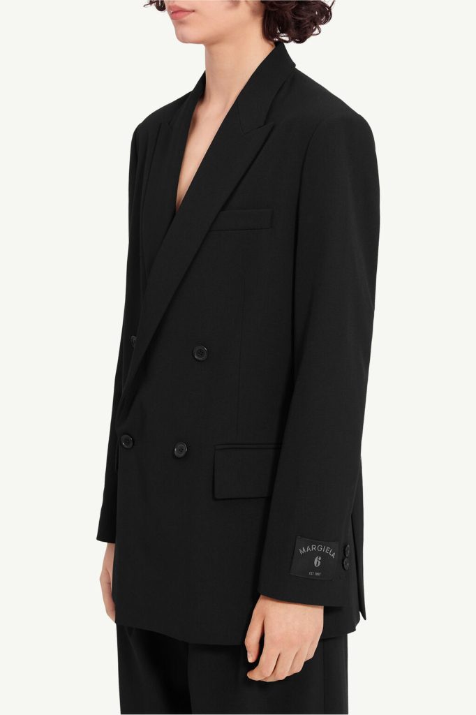Person wearing a black double-breasted blazer with peak lapels; cuff features a Margiela label reading 'Margiela 6 EST 1987'